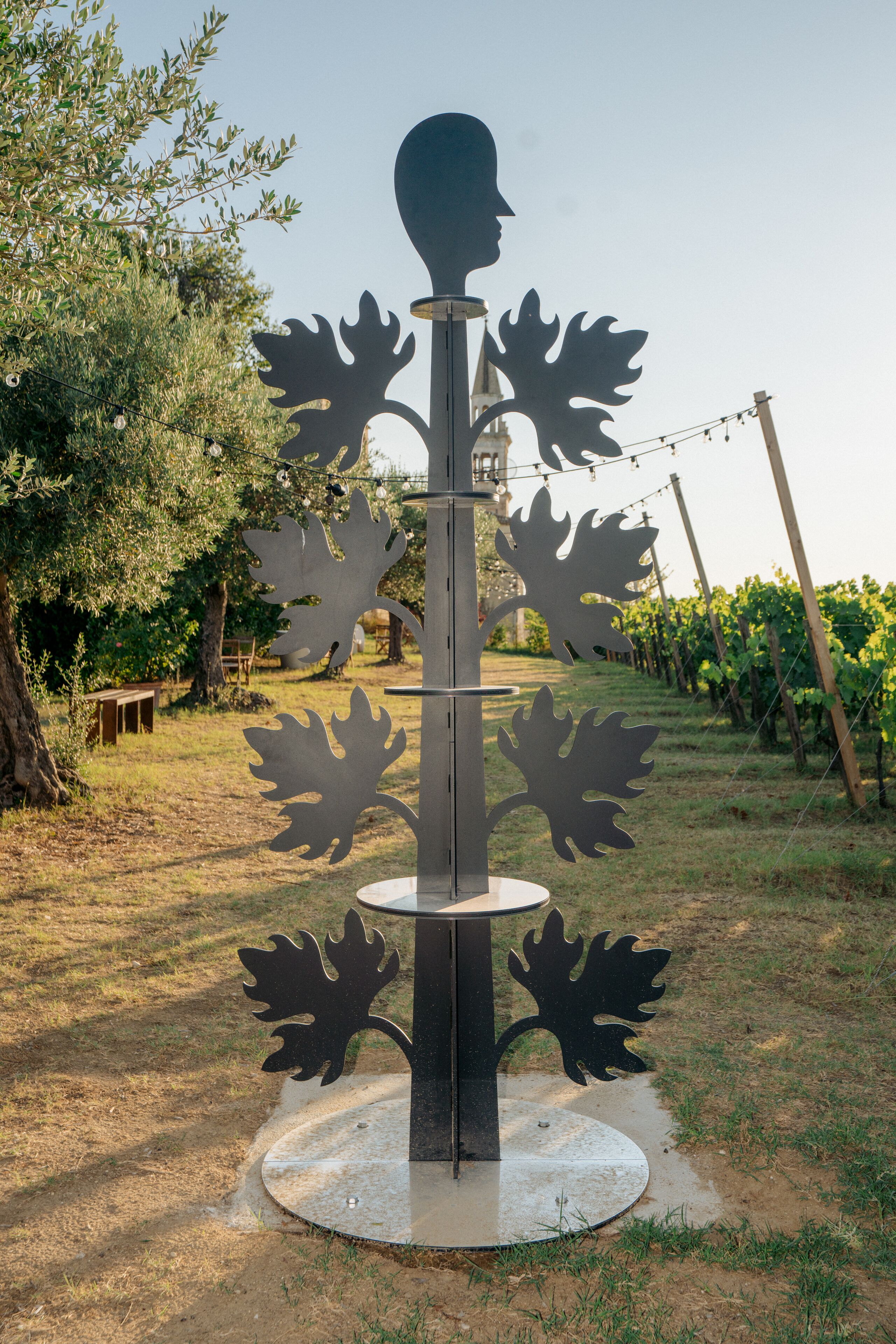 A sculpture with a human profile and leaf-like structures, set on a circular base in a grassy area with trees and a vineyard in the background.