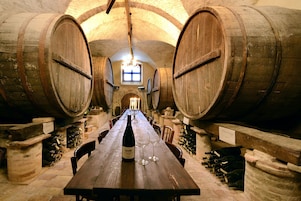 Una larga mesa de madera con una botella de vino y copas en el centro, flanqueada por pilares de piedra y barriles de madera.