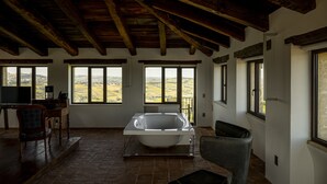 Une pièce spacieuse dotée d’une grande baignoire, de poutres apparentes et de larges fenêtres offrant une vue sur la campagne.