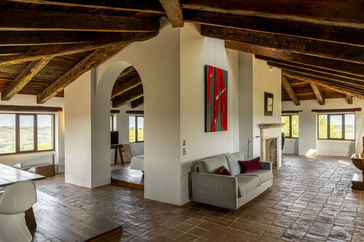 A spacious living area with a high ceiling, exposed wooden beams, a fireplace, and large windows offering a view of the outdoors.