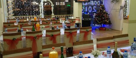 Un restaurante con mesas dispuestas para una comida, sillas adornadas con lazos rojos, un árbol de Navidad y una pantalla de televisión mostrando una chimenea.