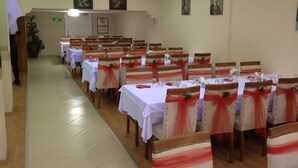 A banquet hall with rows of tables, each with a white tablecloth and red sashes. Chairs are upholstered in a light color with red sashes. The room has a tiled floor and a ceiling with recessed lighting. - Ayvazali Hotel (Bergama)