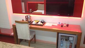 A hotel room with a wooden desk, a chair, a small refrigerator, a television, and a mirror.