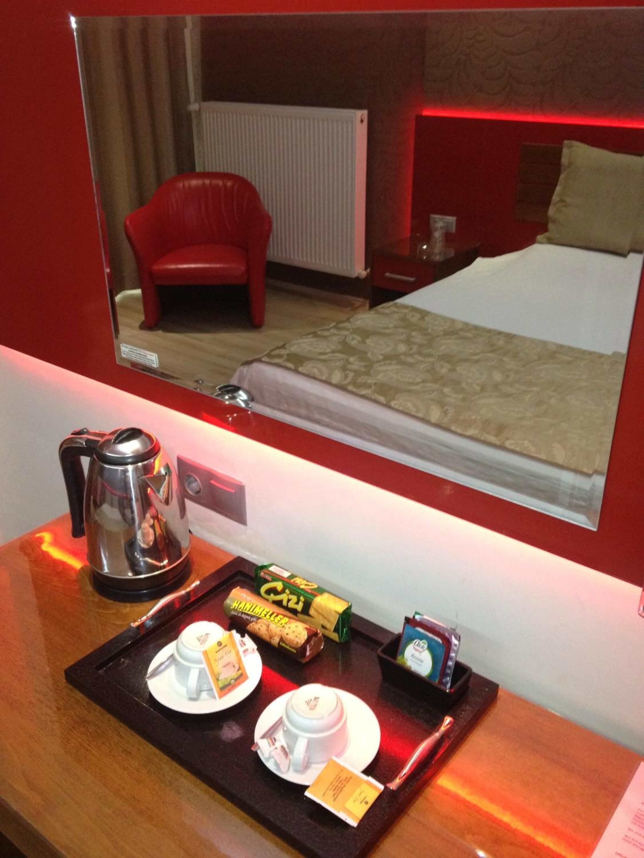 Habitación de hotel con cama, una silla roja, mesita de noche con hervidor, bandeja con artículos de desayuno y un espejo que refleja la habitación.