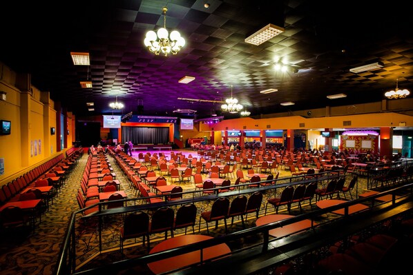 A large banquet hall with rows of tables and chairs, a stage, and a ceiling with patterned tiles and chandeliers.