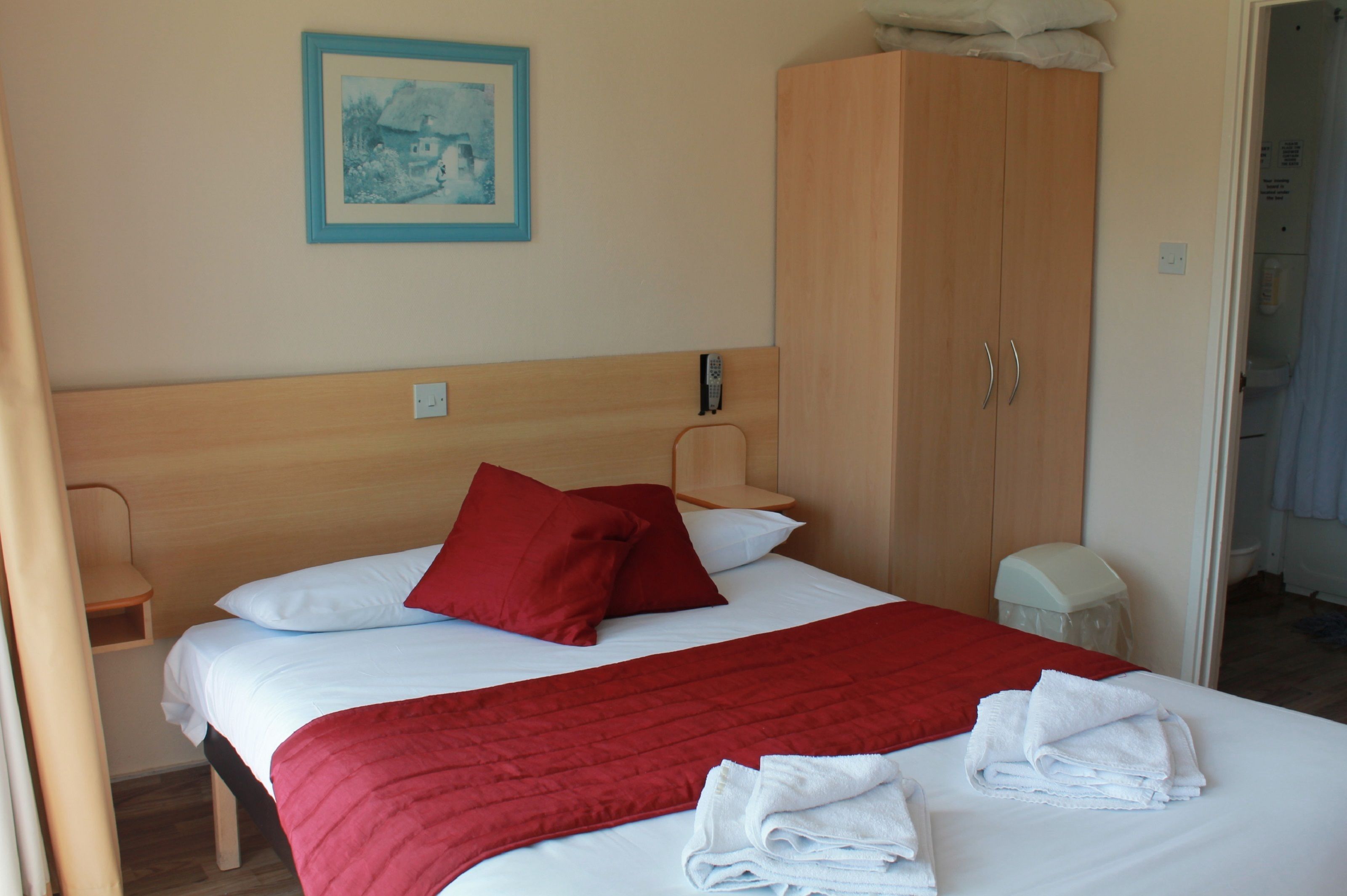 A hotel room with a bed, red pillows, white linens, a red blanket, and a wooden wardrobe.
