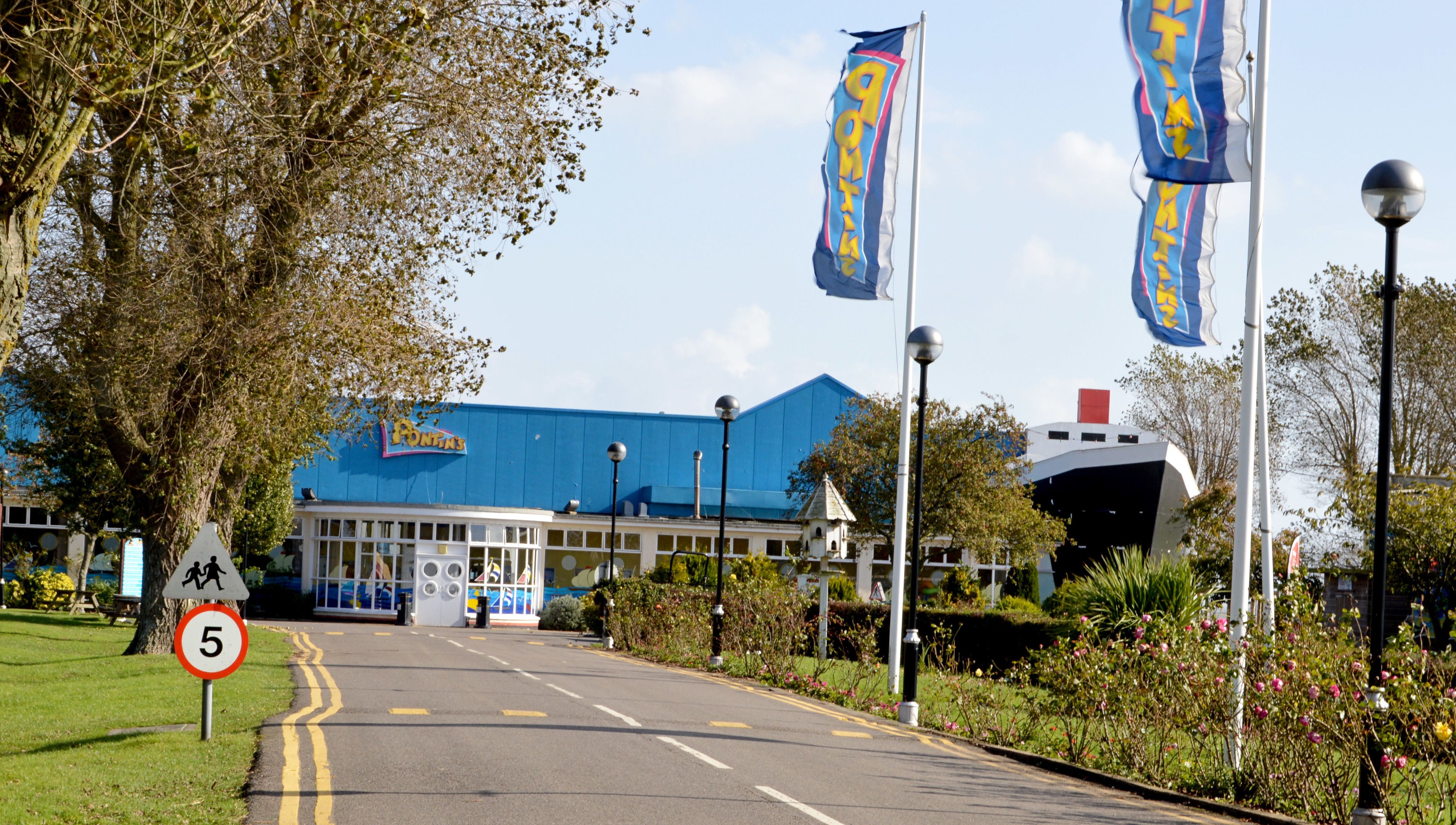 A road leading to a building with a blue roof, surrounded by greenery and flags.