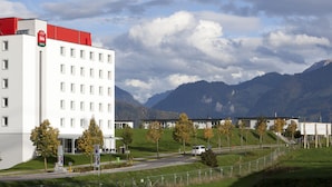 A modern hotel with multiple floors, a prominent red and white logo, and a view of mountains in the background. - ibis Bulle La Gruyere (Bulle)