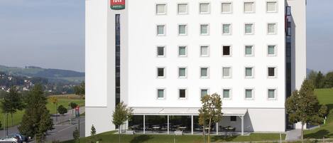 Un hotel moderno con fachada blanca y detalles rojos, que cuenta con varios balcones y una zona de estar exterior cubierta.