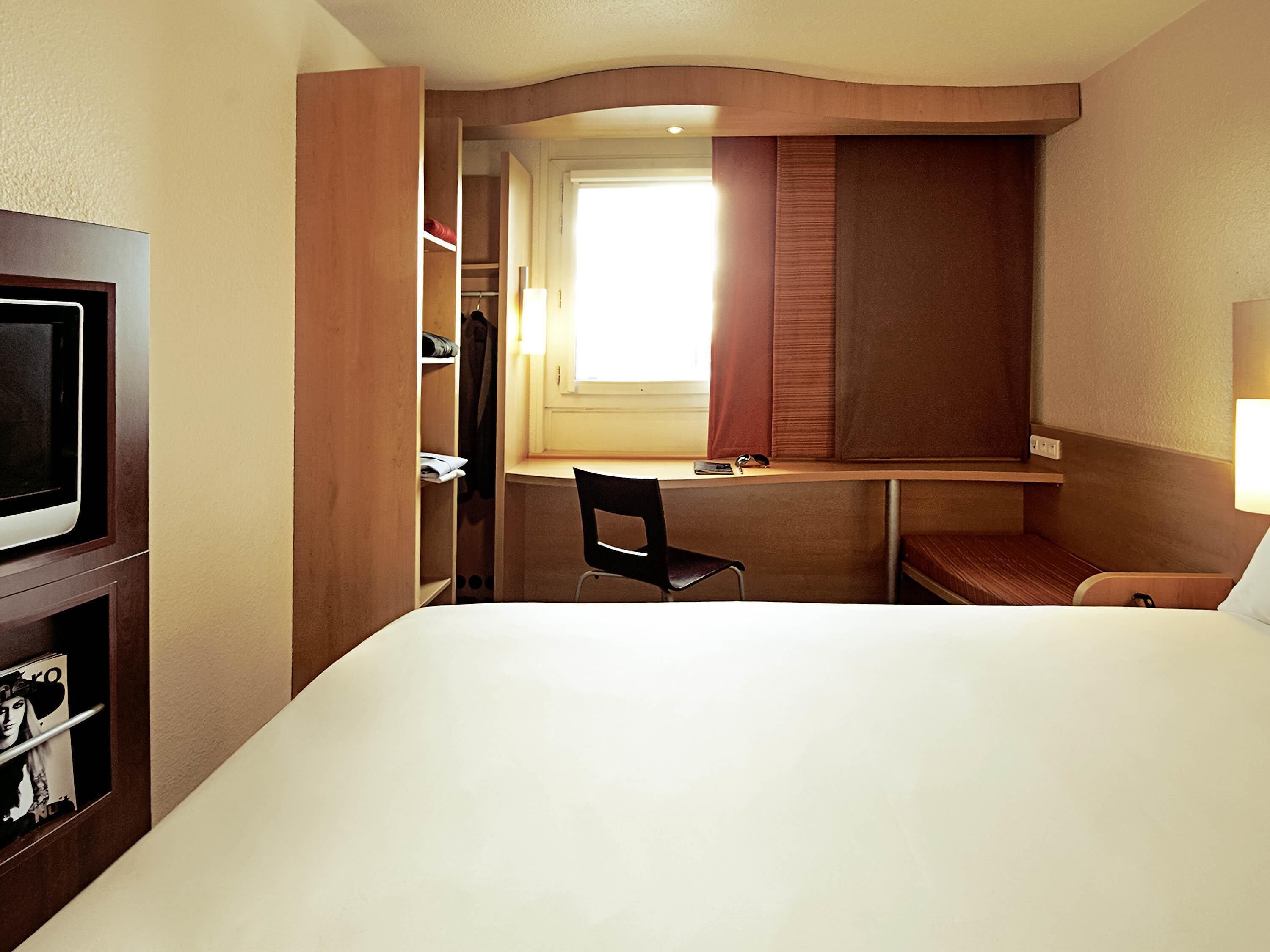 A hotel room with a bed, a desk, a chair, a TV, and a wardrobe.