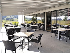 Un café al aire libre con mesas y sillas, ventanas amplias y vistas al campo.