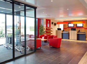 A hotel lobby with a reception desk, red chairs, a Christmas tree, and a glass wall with a shopping cart. - ibis Bulle La Gruyere (Bulle)