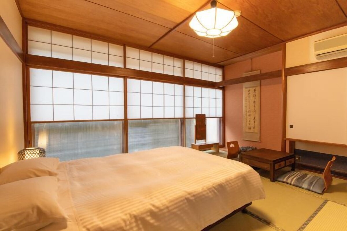 Una habitación tradicional japonesa con suelo de tatami, un gran ventanal de shoji, una cama con cubrecama blanco, un escritorio de madera y un pergamino colgado en la pared.