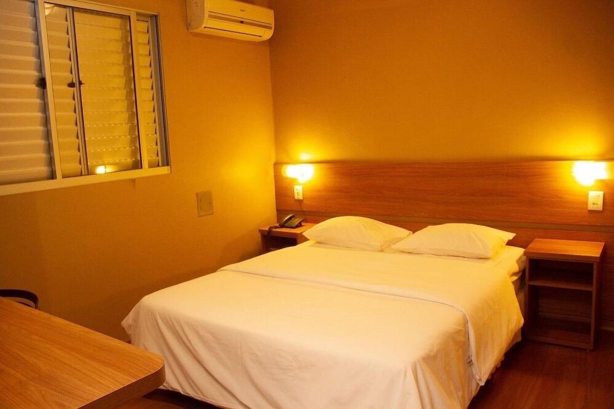 A hotel room with a bed, bedside tables, a desk, and a window with blinds.