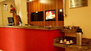 A hotel reception area with a red counter, a coffee machine, a vase with flowers, and a sign listing room rates. - Bernal Hotel (Ituiutaba)