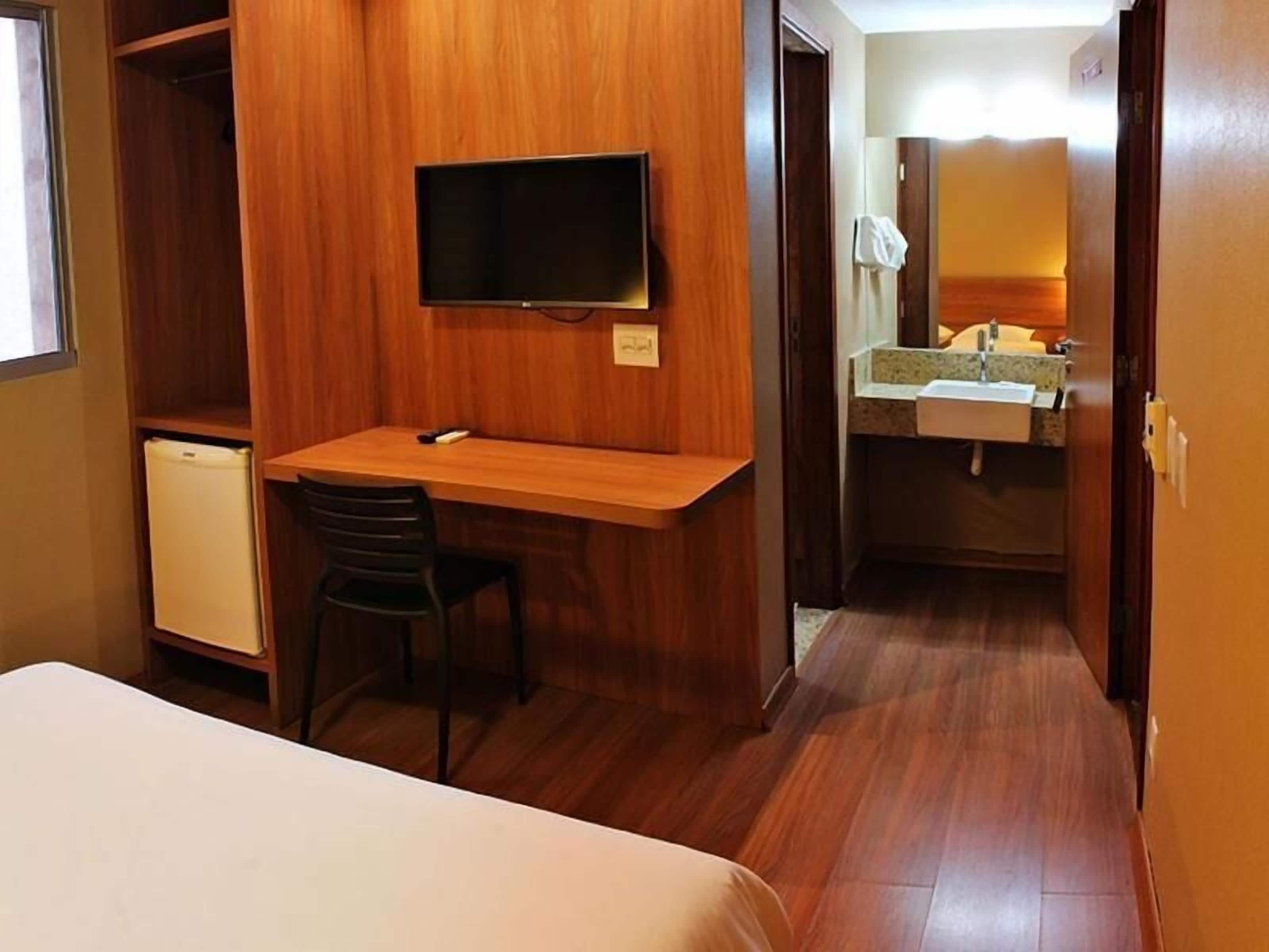 A compact hotel room with a bed, a small desk with a chair, a television, a mini-fridge, and a bathroom with a sink and mirror.