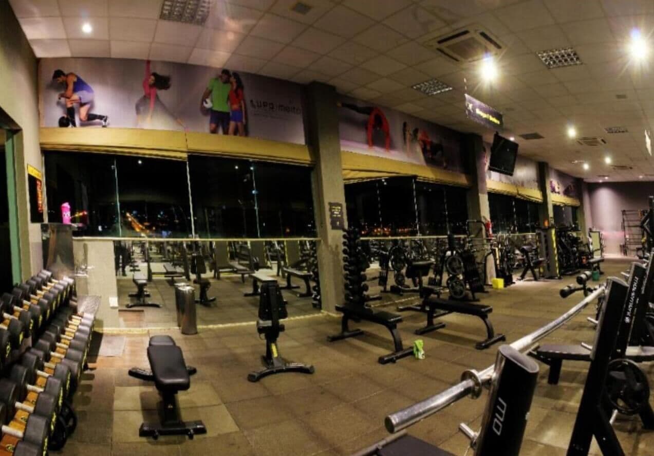 Un gimnasio con diversos equipos de ejercicio, espejos y una pared con anuncios.