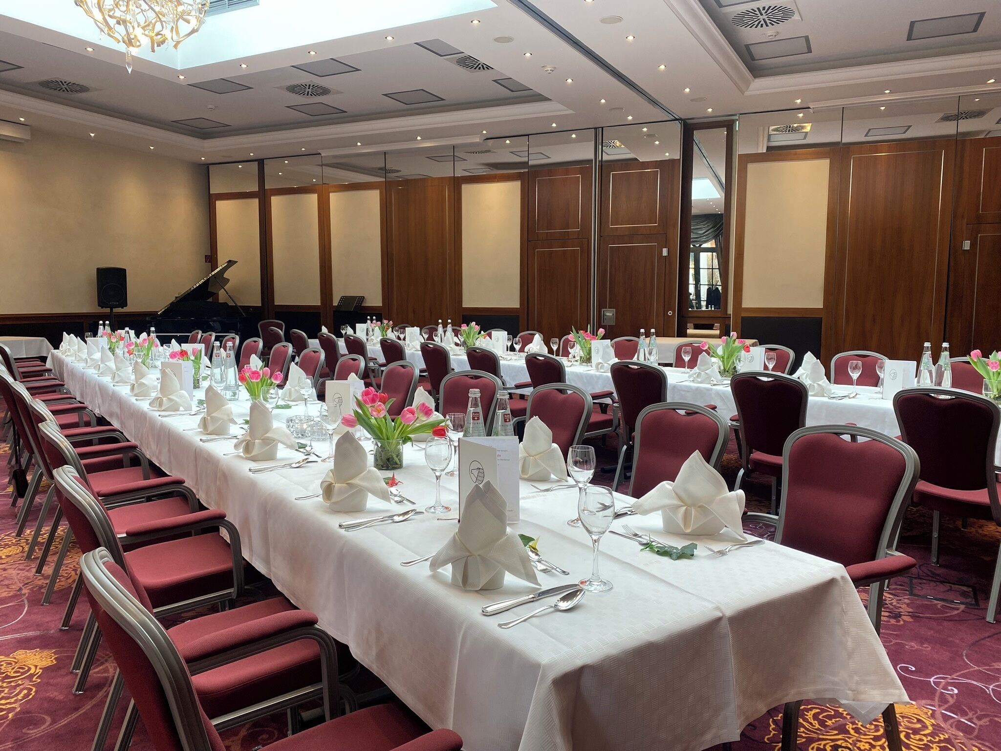 Ein Festsaal mit langen Tischen, roten Stühlen, weißen Tischdecken und floralen Mittelstücken.