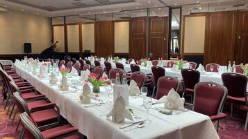 Ein Festsaal mit langen Tischen, roten Stühlen, weißen Tischdecken und floralen Mittelstücken.