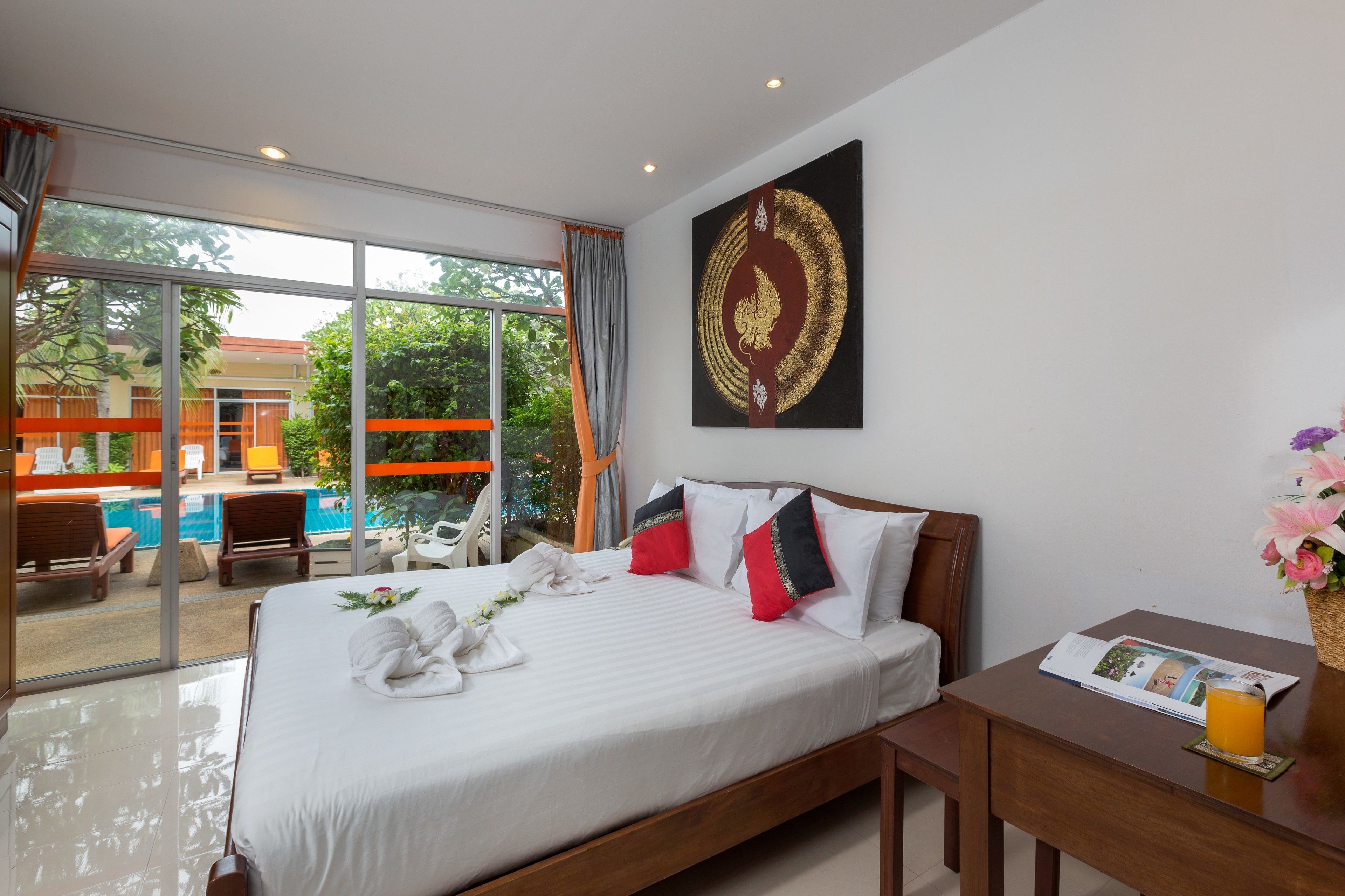 A hotel room with a large bed, a wooden headboard, a bedside table with a magazine and a glass of orange juice, and a large window offering a view of a pool and outdoor seating area.
