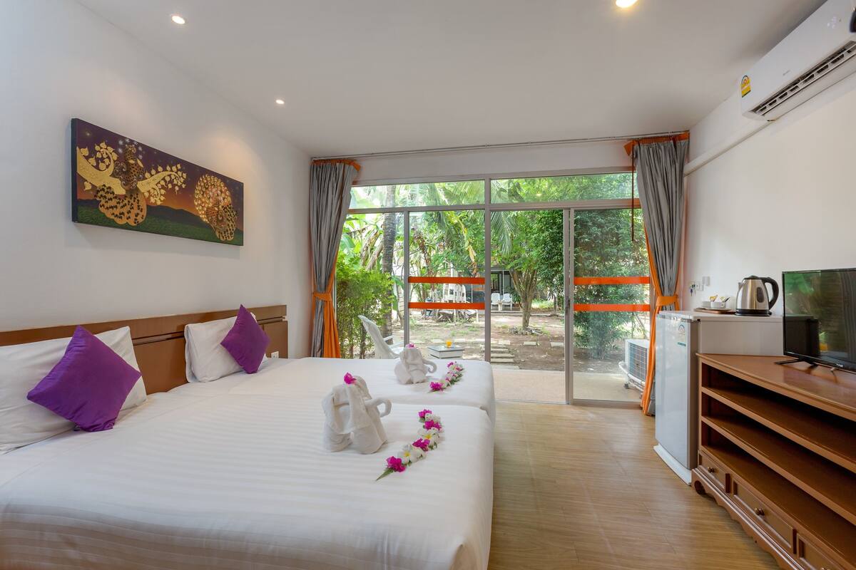 A hotel room with two beds, a TV, a mini-fridge, and a balcony with a view of greenery.