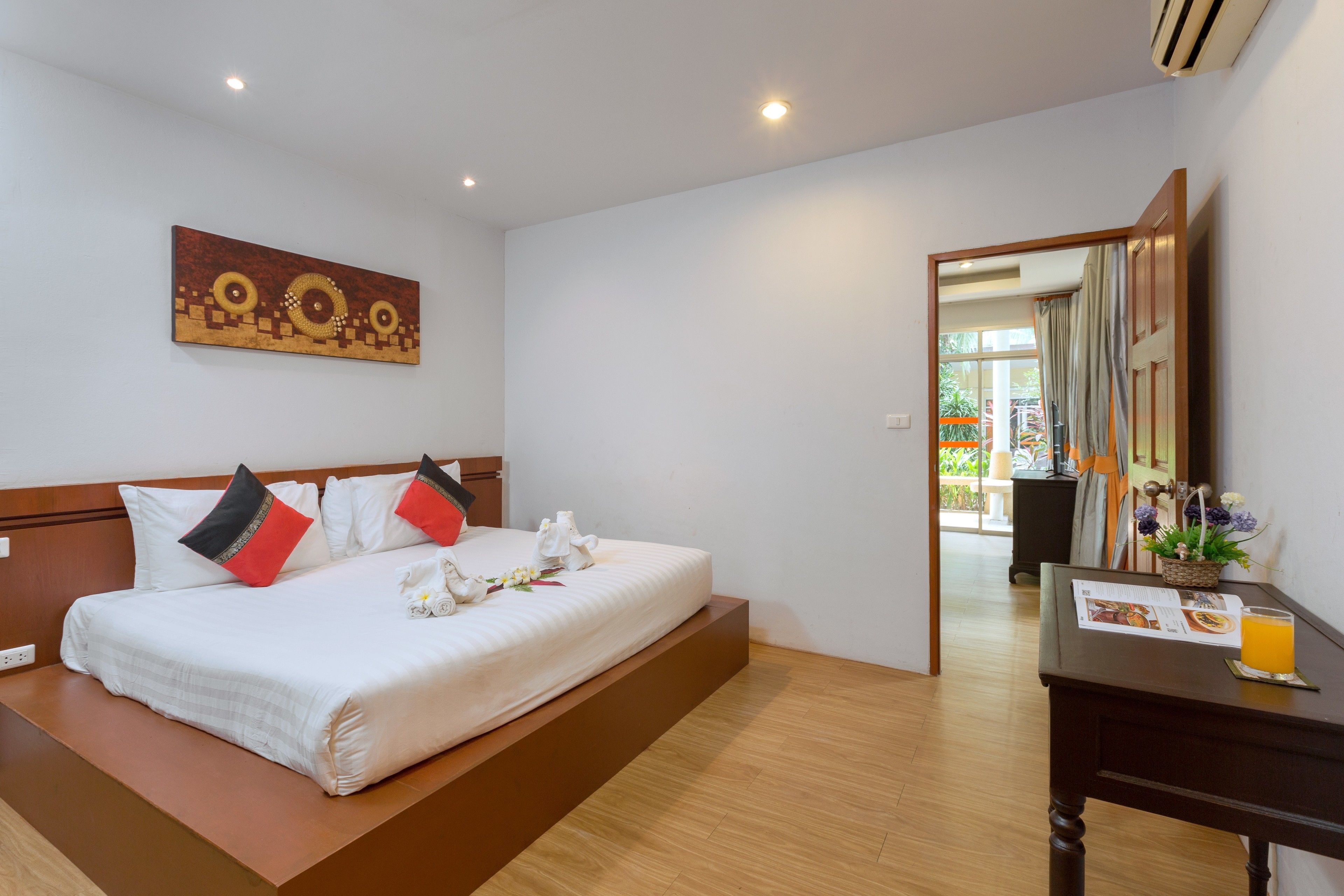 Ein Hotelzimmer mit einem großen Bett, einem hölzernen Kopfteil, einem Bild an der Wand, einem Schreibtisch mit einer Speisekarte und einem Glas Orangensaft sowie Blick durch eine offene Tür.