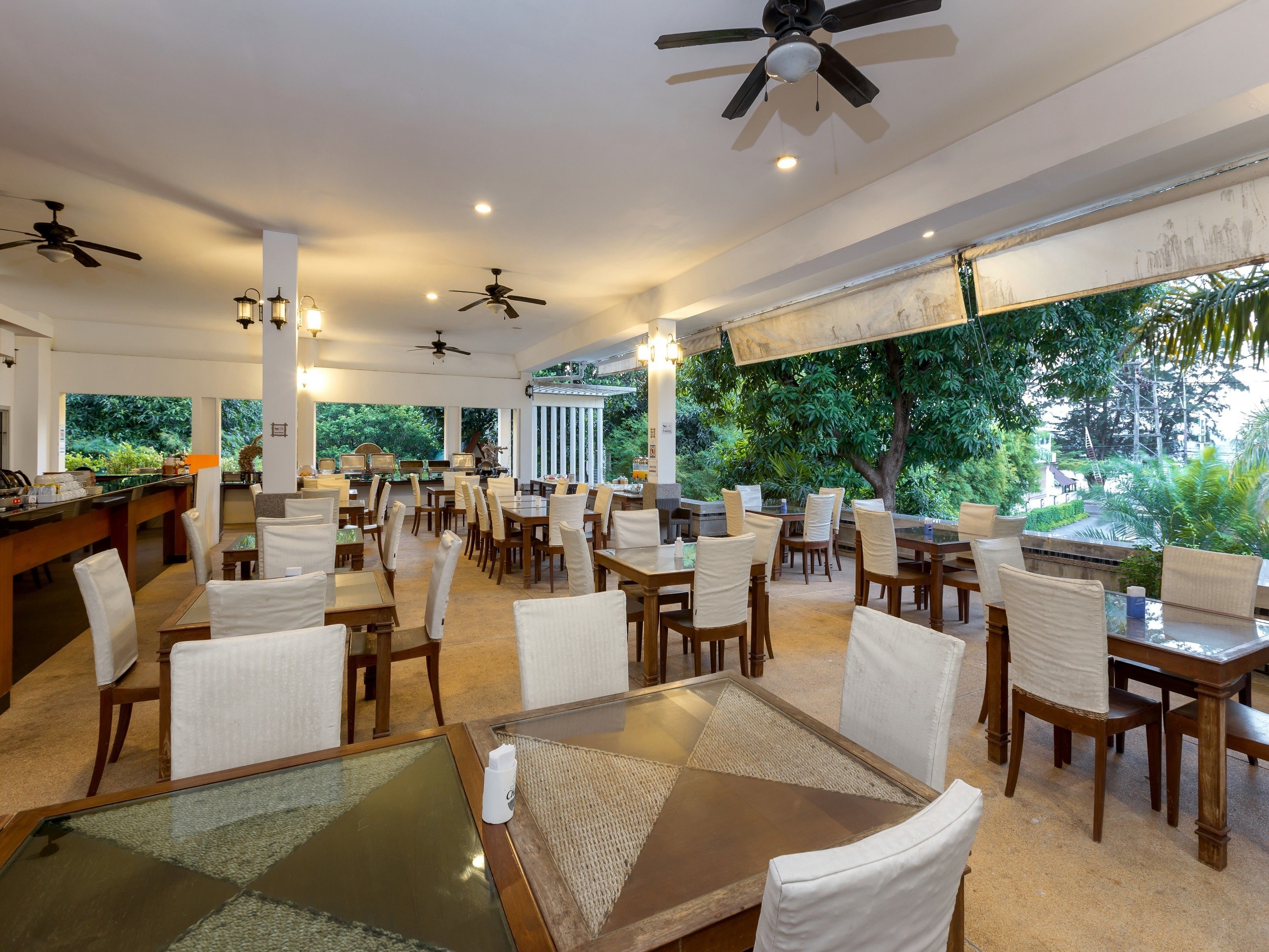 A spacious indoor dining area with multiple tables and chairs, ceiling fans, and large windows offering a view of lush greenery.