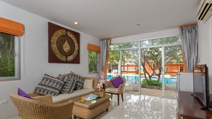 A spacious living area with a sofa, chairs, a coffee table, and a TV. Large windows offer a view of a swimming pool and greenery outside. - Phuket Sea Resort (Rawai)