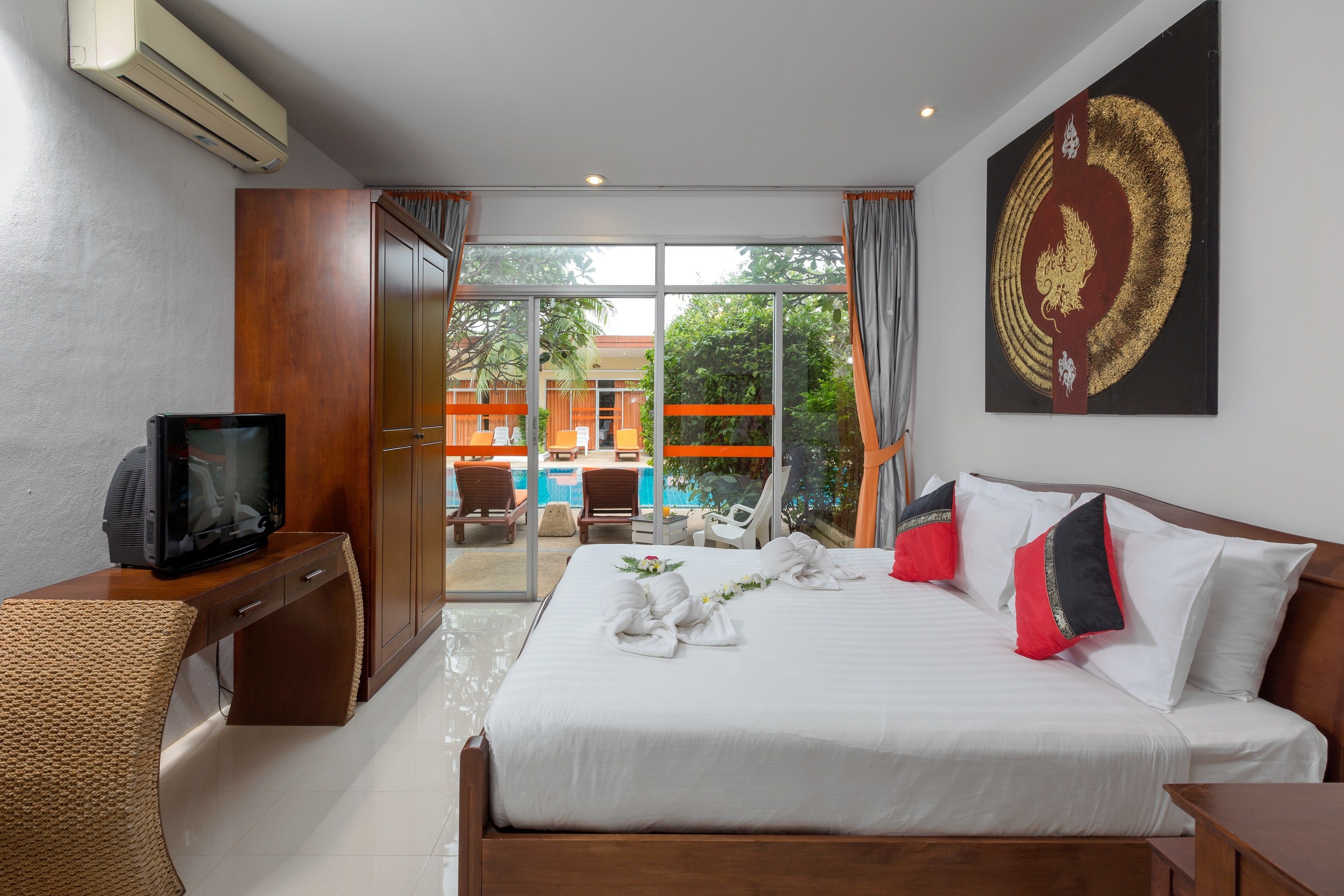Una habitación de hotel con cama, un escritorio con televisor, un ventanal con vista a una piscina y zona de estar al aire libre, y un elemento decorativo en la pared.