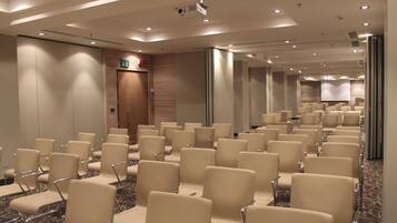 Una sala conferenze con file di sedie beige, un proiettore e una porta con la segnaletica di uscita di sicurezza.