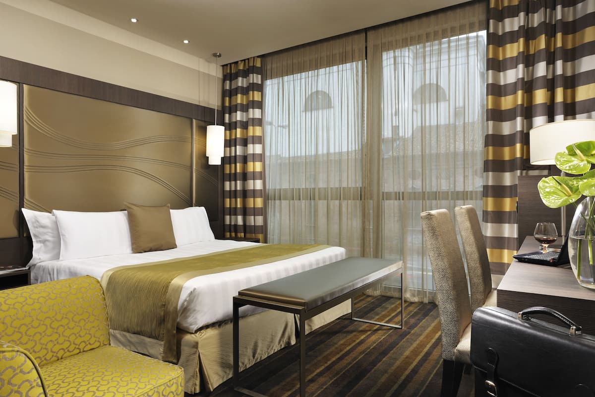 A hotel room with a bed, a bench, a chair, a desk with a laptop, and a plant.