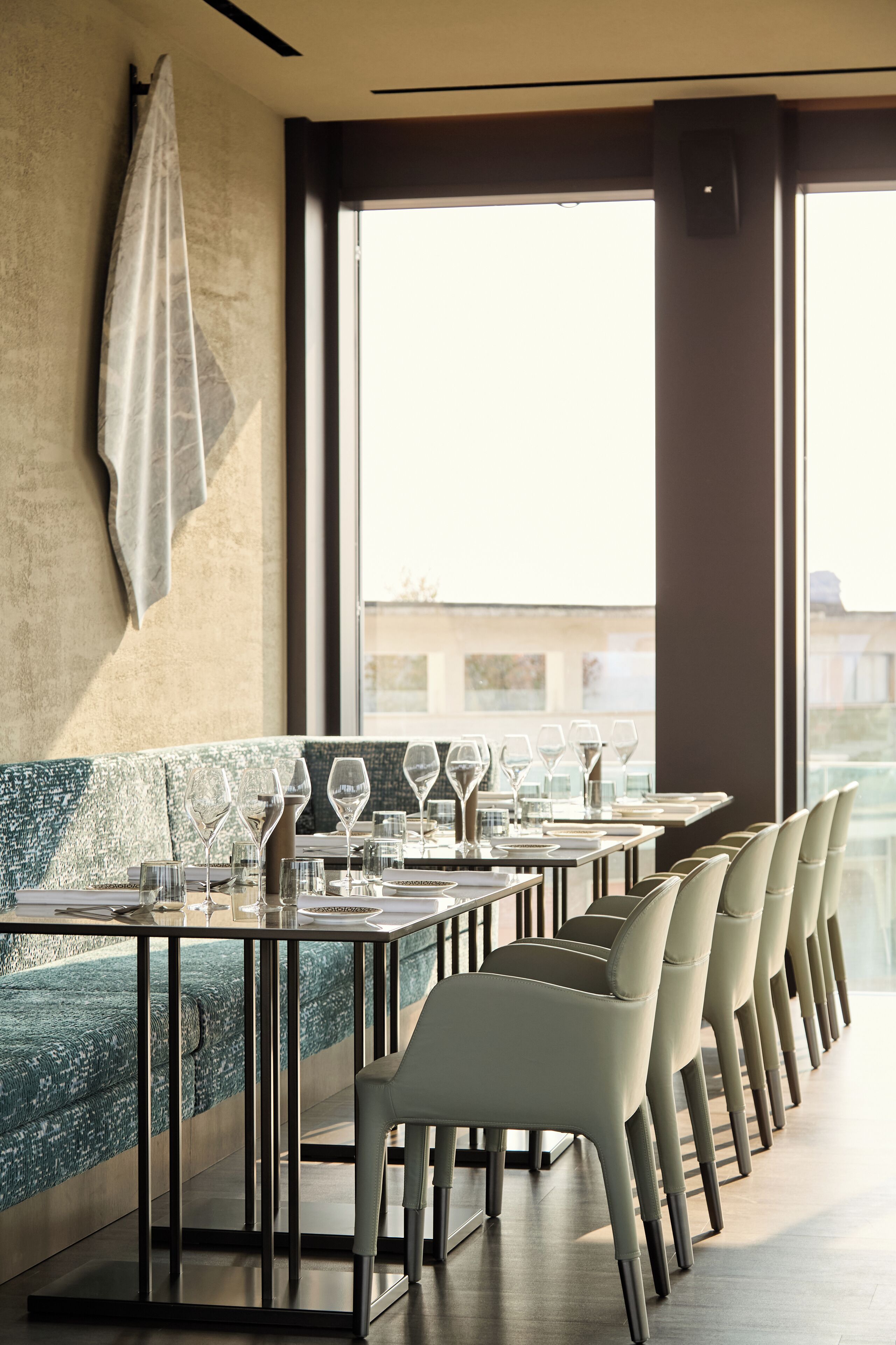 A restaurant with a long table set for dining, chairs aligned along the wall, and a view through large windows.