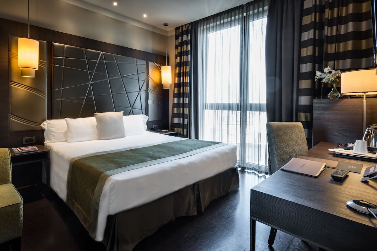 A hotel room with a large bed, a desk with a chair, a nightstand, a wall-mounted lamp, and a window with curtains.