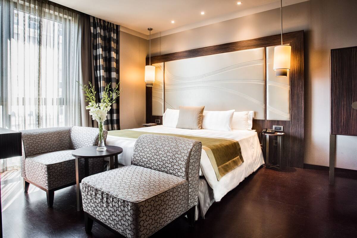 A hotel room with a bed, two armchairs, a small table, a vase with flowers, and a large window with curtains.