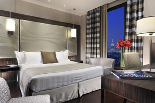 Una camera d'albergo con un letto grande, una scrivania con una rivista, una sedia e vista sulla città dalla finestra.