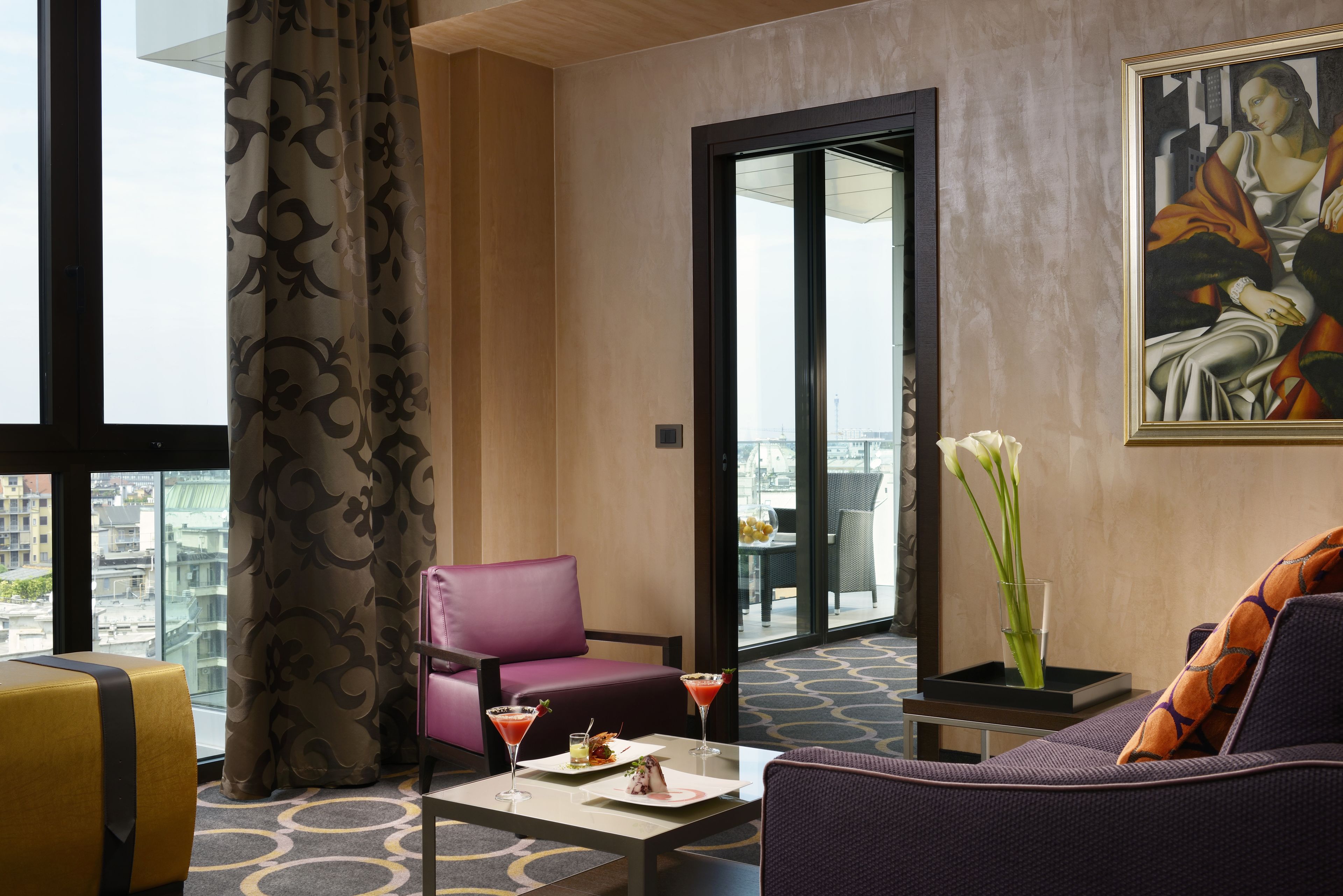 A hotel room with a large window, a purple sofa, a pink chair, a small table with a martini, and a vase with flowers.