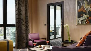 A hotel room with a large window, a purple sofa, a pink chair, a small table with a martini, and a vase with flowers.