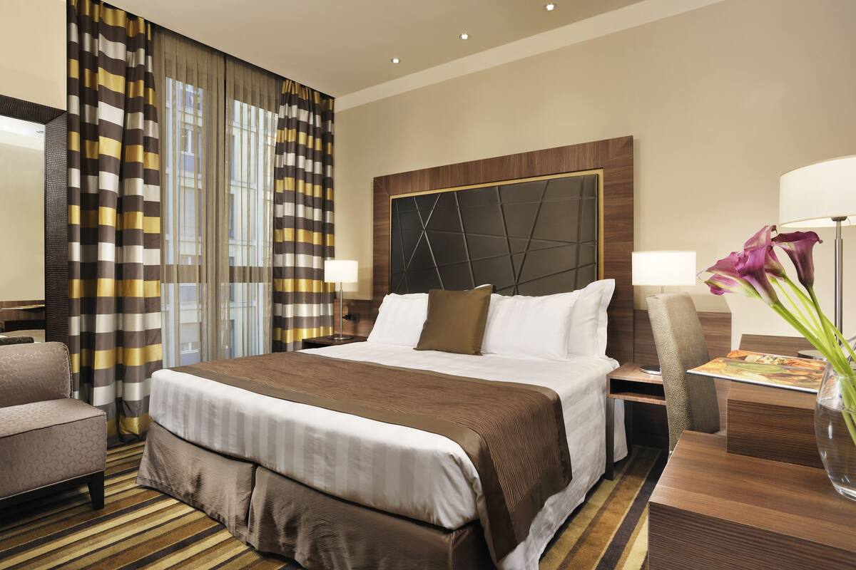 A hotel room with a large bed, a desk, a chair, a vase with flowers, and a window with curtains.