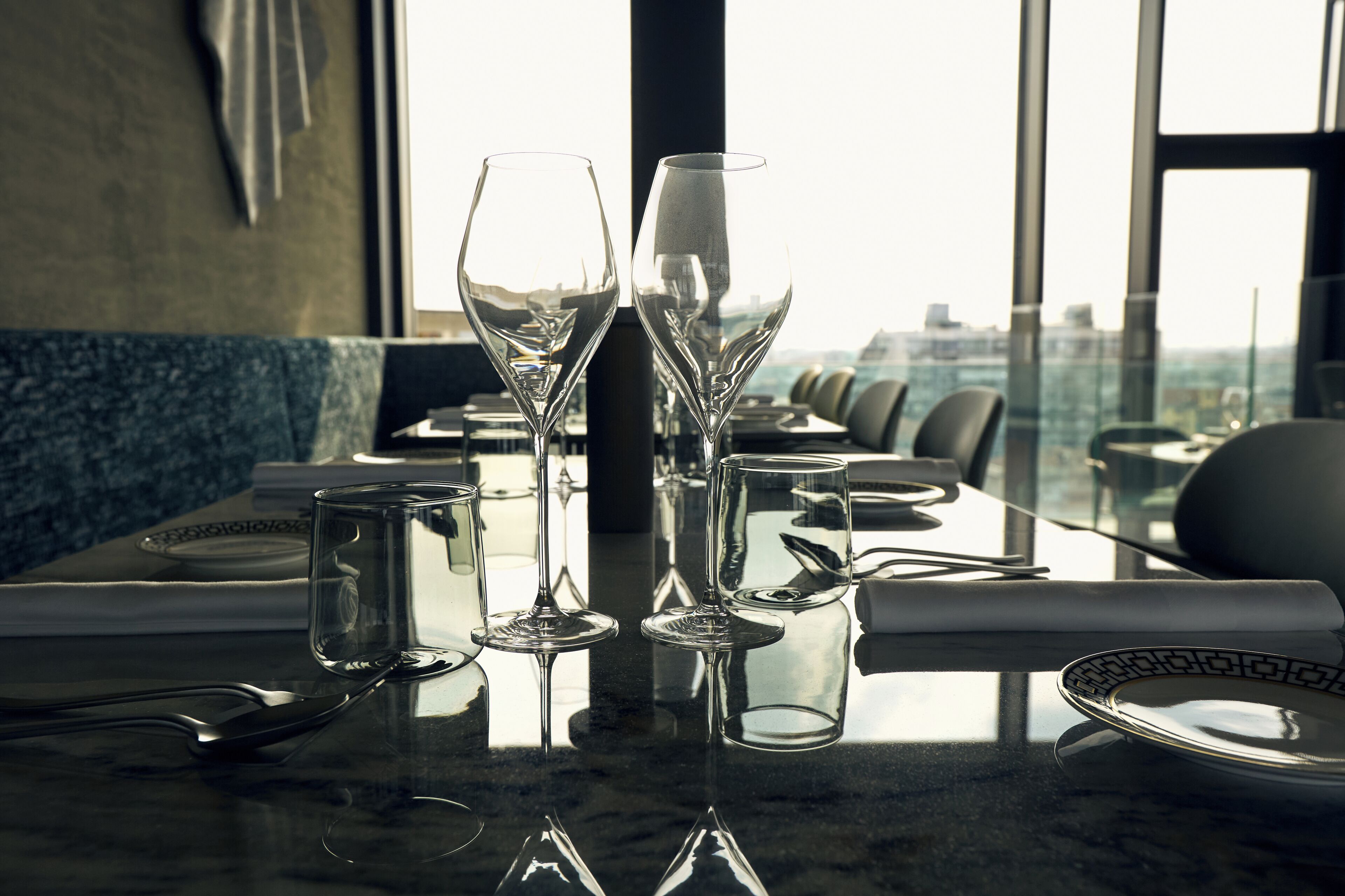 Una mesa de comedor con copas y platos vacíos, en un restaurante moderno con vista a la ciudad.