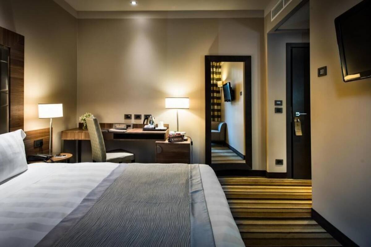 Una camera d'hotel con un letto, una scrivania, una sedia, una lampada e un televisore fissato alla parete.
