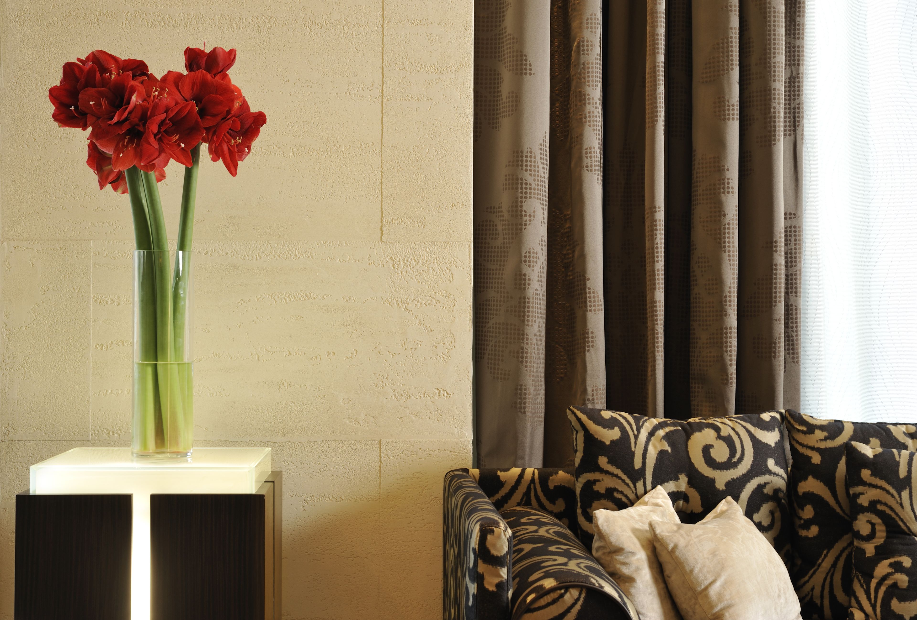 A living room with a patterned sofa, a vase with red flowers on a side table, and a window with curtains.