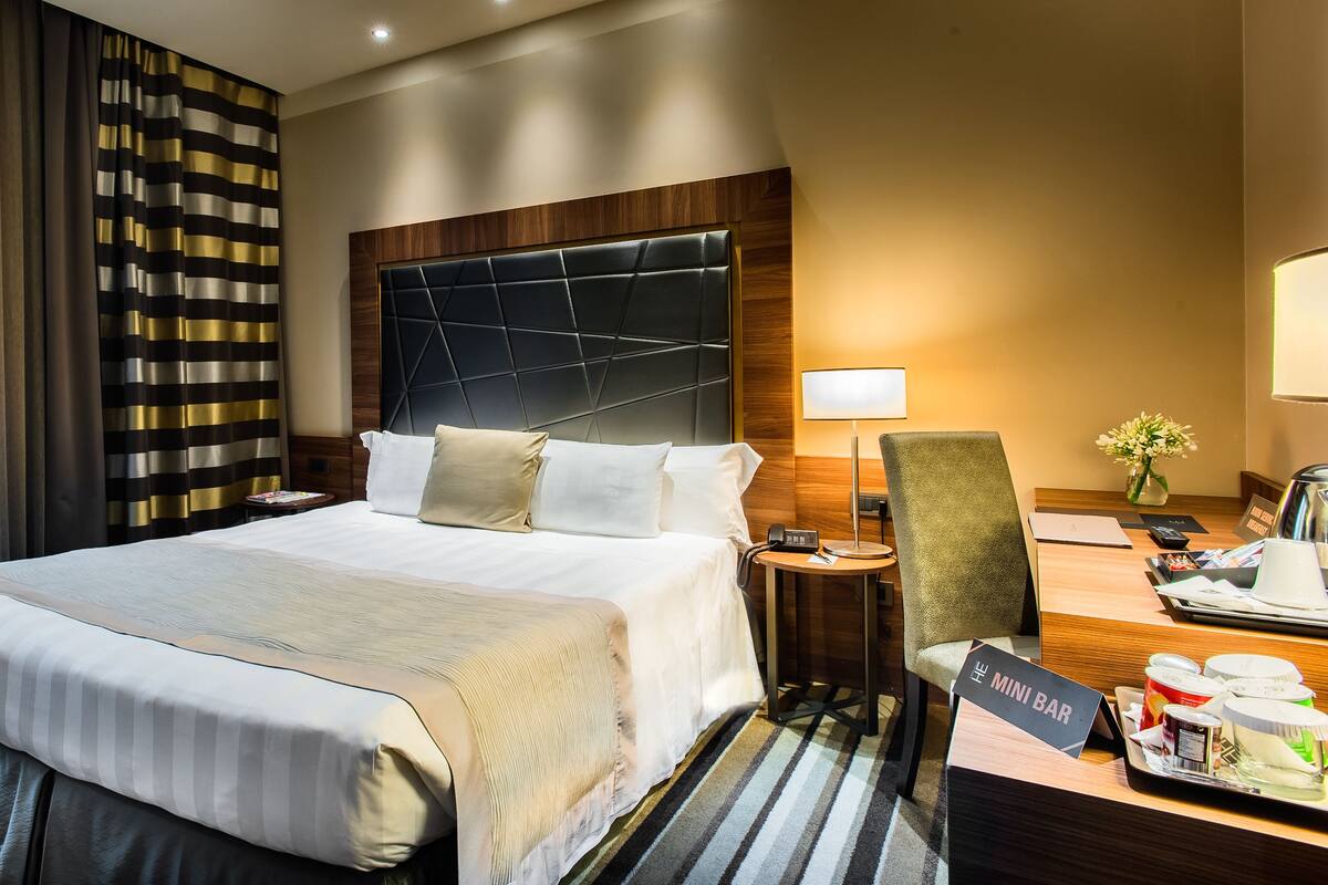 A hotel room with a large bed, a bedside table with a lamp, a chair, a mini bar, and a view of the outdoors.