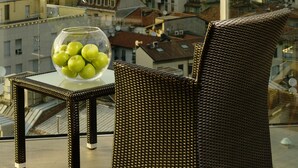 A balcony with a wicker chair and a glass table holding a bowl of apples, overlooking a cityscape with historic architecture and modern buildings.
