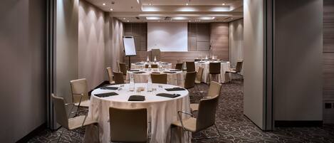 A conference room with multiple round tables, chairs, a projector screen, and a presentation screen.