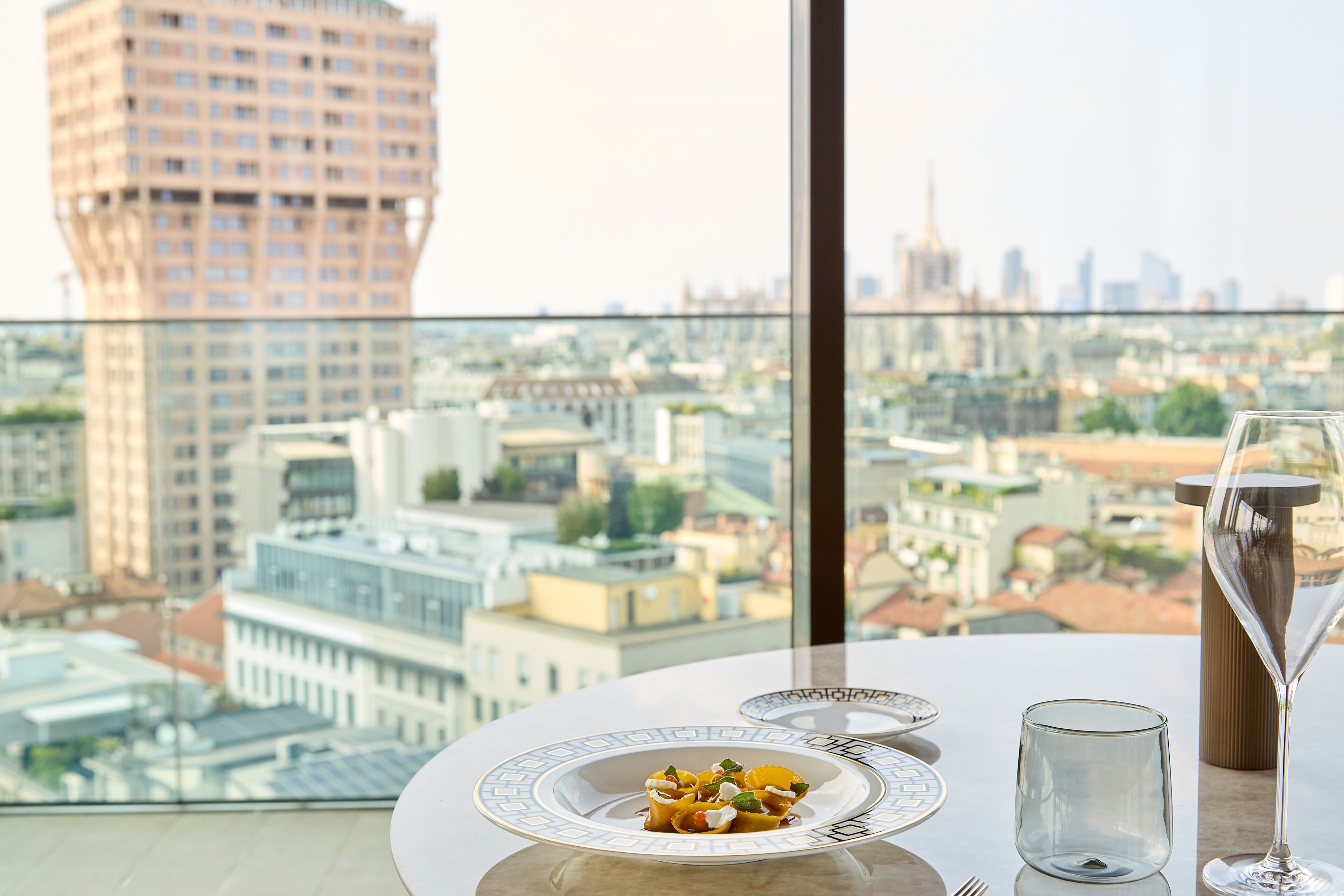A dining table with a plate of food and a wine glass, overlooking a cityscape with buildings.