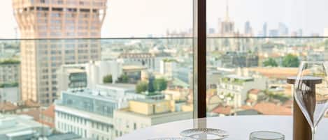Ein Esstisch mit einem Teller Essen und einem Weinglas, mit Blick auf eine Stadtlandschaft und Gebäude.