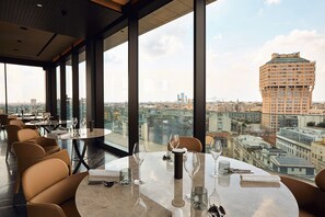 A restaurant with a panoramic city view, featuring round tables and modern chairs.