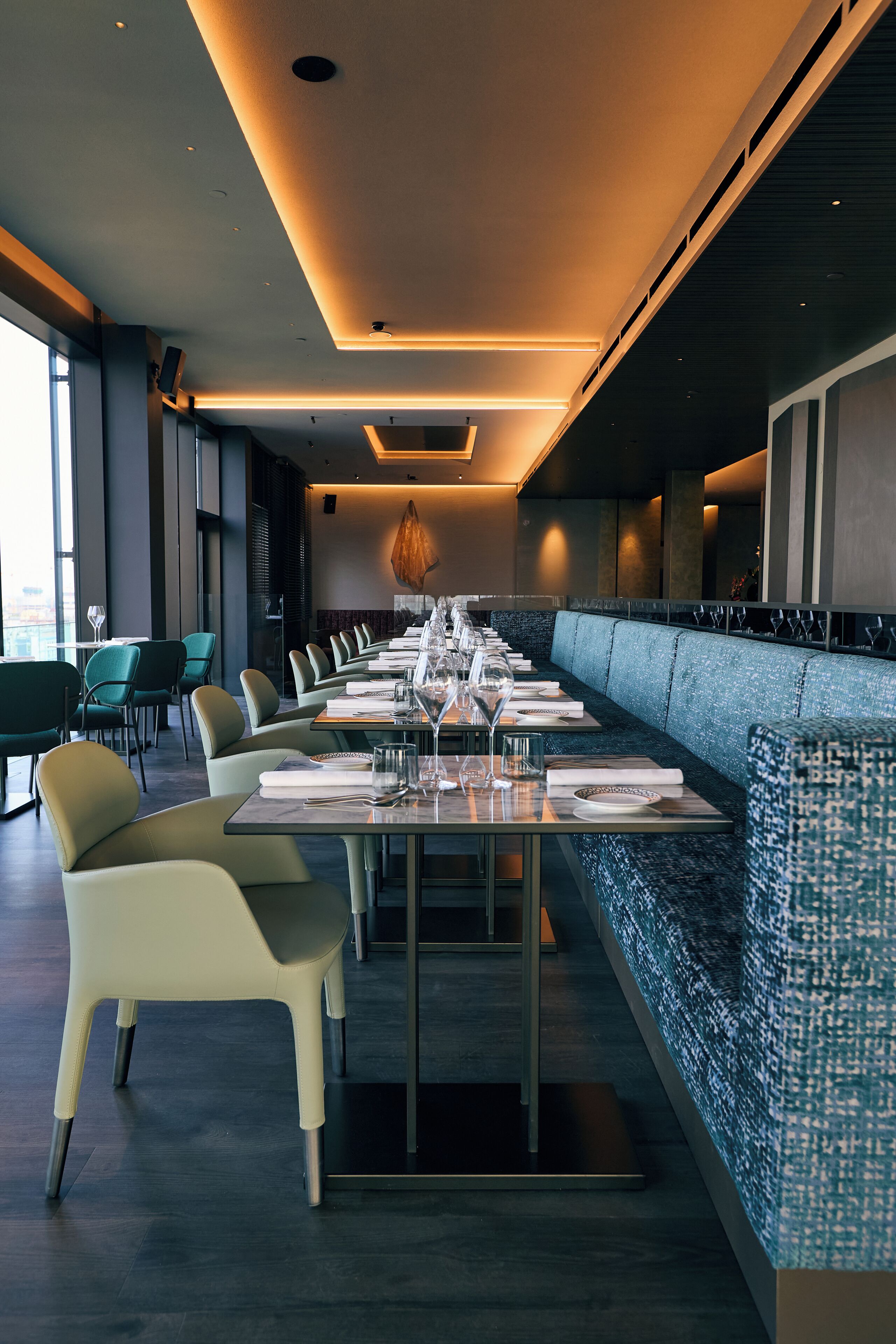 A modern restaurant with a long dining table set for a meal, surrounded by cushioned seating.