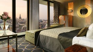 A hotel room with a large bed, a view of the cityscape, a round mirror, and a vase with flowers on a table.