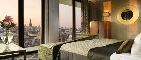 Habitación de hotel con una cama grande, vista panorámica de la ciudad, un espejo redondo y un jarrón con flores sobre una mesa.